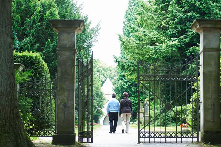 Paar geht durch ein offenes schmiedeeisernes Tor in einen Friedhof mit einem Blick auf eine Kapelle.