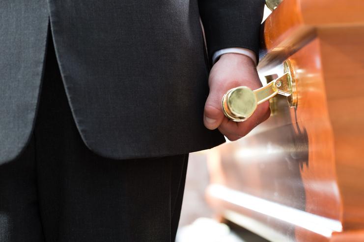 Mann in Anzug hält eine goldene Griffschleife an einem Holzsarg.