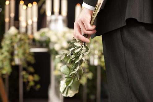 Mann in schwarzem Anzug hält einen Strauß mit grünen Blättern und einer weißen Rose vor einem dekorierten Hintergrund.