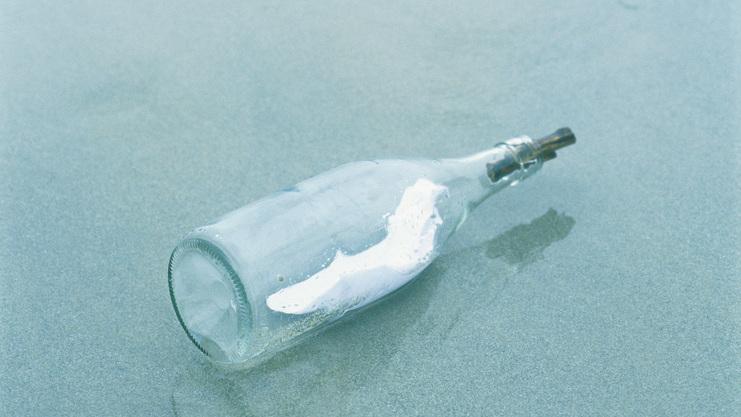 Eine Glasflasche liegt am Strand, umgeben von sanften Wellen und feinem Sand. In der Flasche ist ein dünnes Papier.