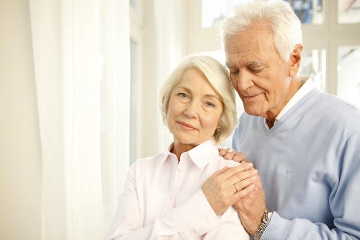 Ein älteres Paar steht nah beieinander, der Mann umarmt die Frau sanft, beide lächeln in eine ruhige Umgebung.