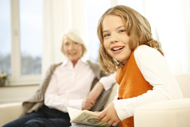 Lächelndes Mädchen mit Buch, im Hintergrund eine ältere Frau, beide in heller Wohnumgebung.